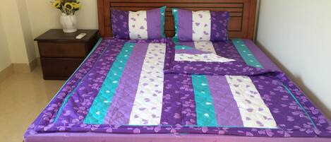 A bed with a purple and teal quilt, a wooden headboard, and a nightstand with a vase of sunflowers.