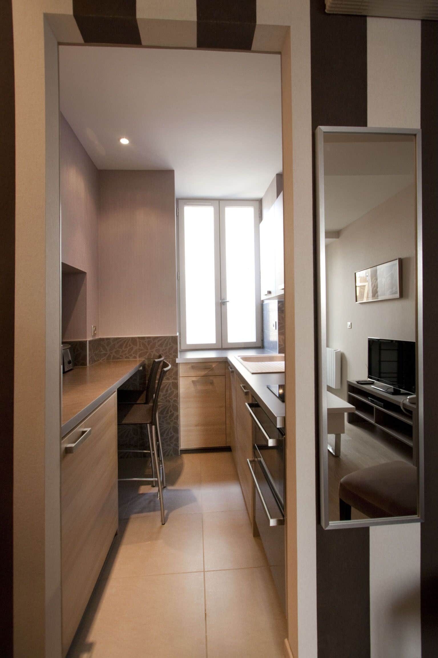 A modern kitchen with a built-in oven, a microwave, and a sink. There is a window providing natural light, and a mirror is mounted on the wall.