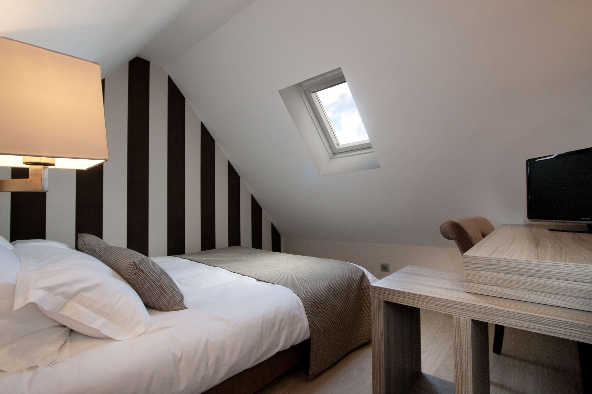 A bedroom with a sloped ceiling, a skylight, a bed with white bedding, a bedside table, and a television.