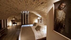 Un dormitorio con techo de piedra, una cama con ropa de cama blanca, una silla de madera, una escalera y un cuadro enmarcado en la pared.
