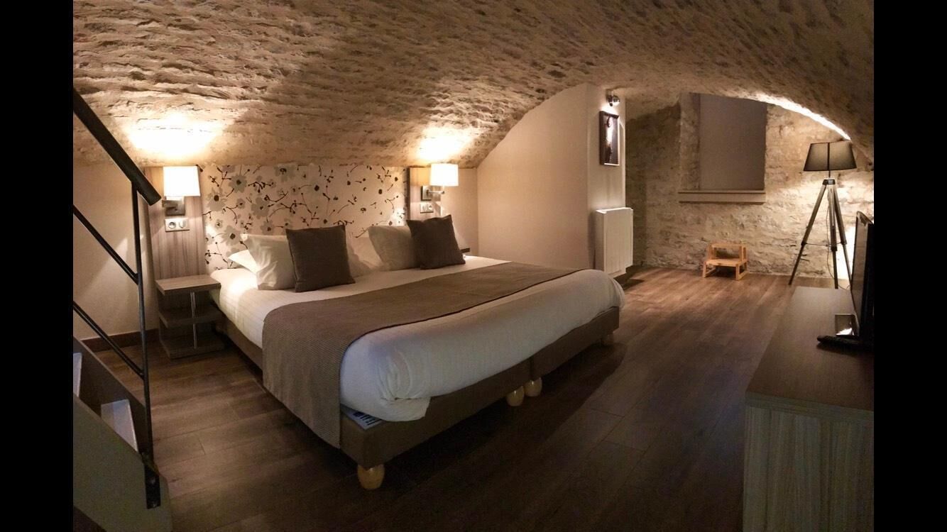 Un dormitorio con techo de piedra, una cama con cubrecama blanco, dos lámparas de noche, una pequeña silla de madera y una lámpara de pie.