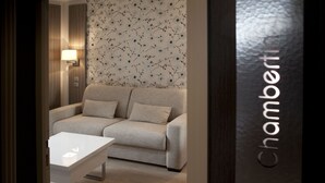 Una habitación de hotel moderna con un sofá, una mesita y una lámpara fijada a la pared. La habitación tiene suelo de madera y papel pintado con motivos florales.