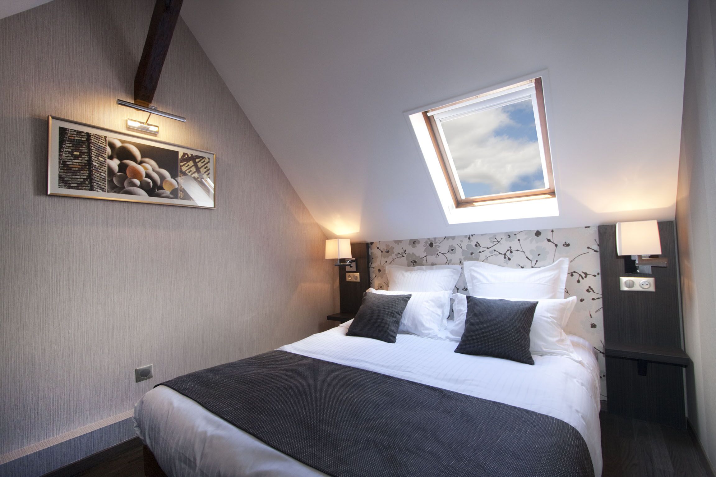Una habitación de hotel moderna con una cama grande, un tragaluz, una obra de arte en la pared y una mesita de noche con lámpara.