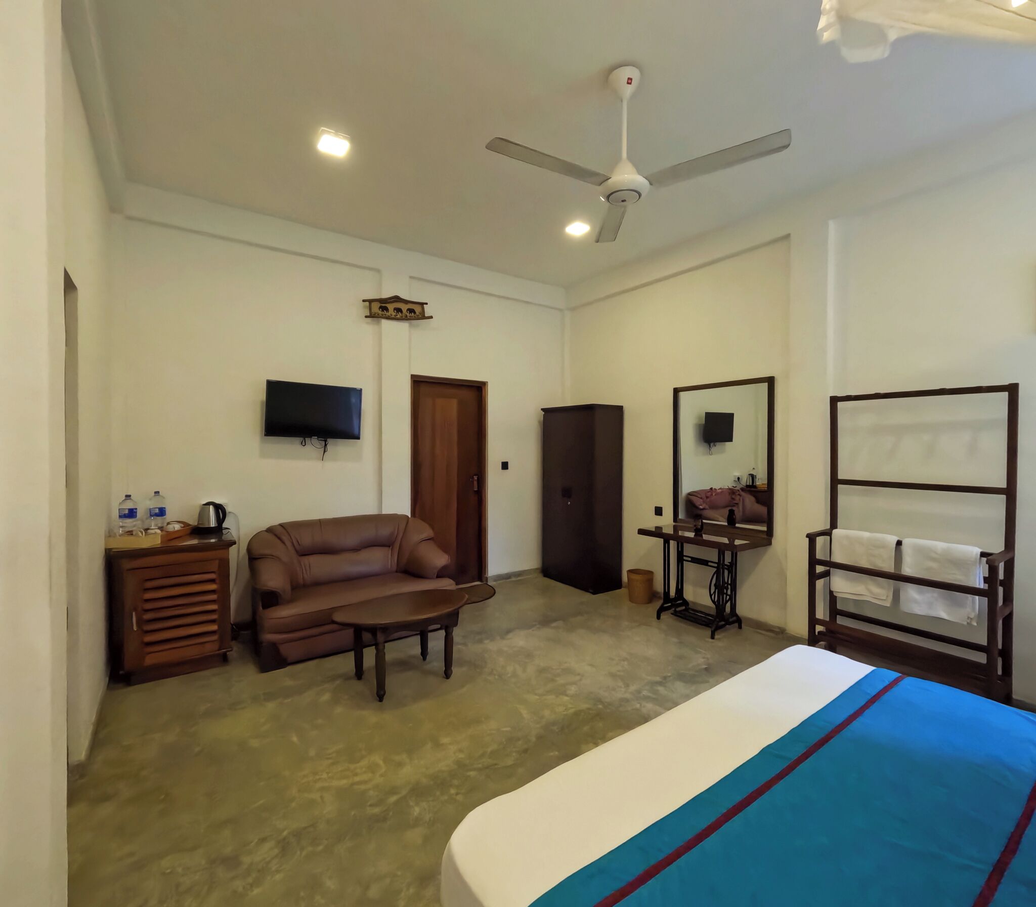 A room with a bed, a sofa, a small table, a television, a ceiling fan, and a mirror.
