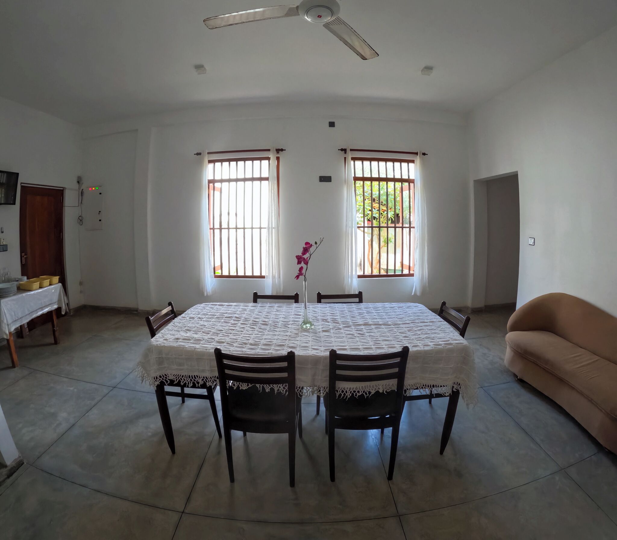 Uma sala de jantar com uma mesa posta para seis pessoas, duas janelas com balaústres, um ventilador de teto e um sofá.