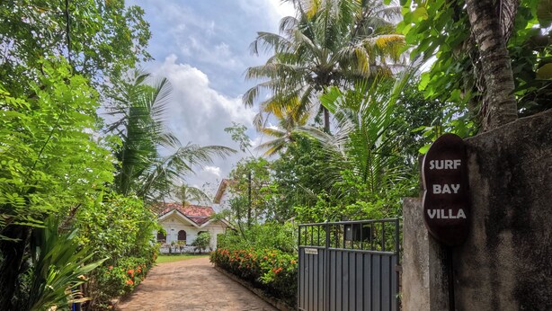A gated entrance to a property named 'Surf Bay Villa' with a stone pathway leading to a building with a red-tiled roof. - Surf Bay Villa (Dikwella)