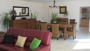 A living room with a red leather sofa, a dining table with wicker chairs, and a kitchen in the background.