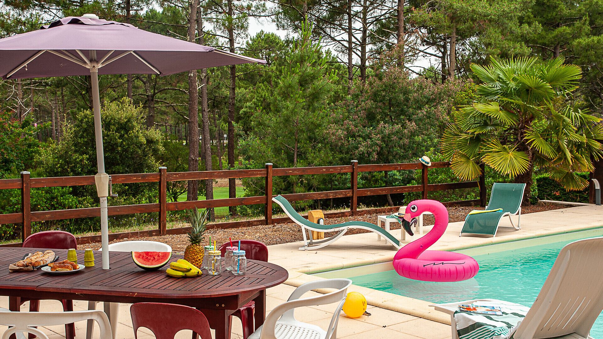 Ett område vid poolen med ett trästaket, solstolar, ett bord med mat och dryck samt en uppblåsbar flamingofigur.