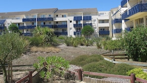 Ein mehrstöckiges Hotel mit Balkonen, ein Gartenbereich mit Sträuchern und einem Holzzaun, sowie ein klarer, blauer Himmel.