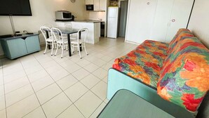 Un espacio habitable compacto con kitchenette, una zona de comedor con sillas blancas, un sofá con una manta colorida y un televisor montado en la pared.