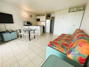 Um espaço compacto com cozinha compacta, área de refeições com cadeiras brancas, sofá com manta colorida e televisão na parede.