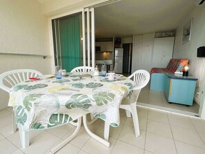 Uma área de refeições com uma mesa coberta por uma toalha com padrão de folhas, cercada por cadeiras brancas.