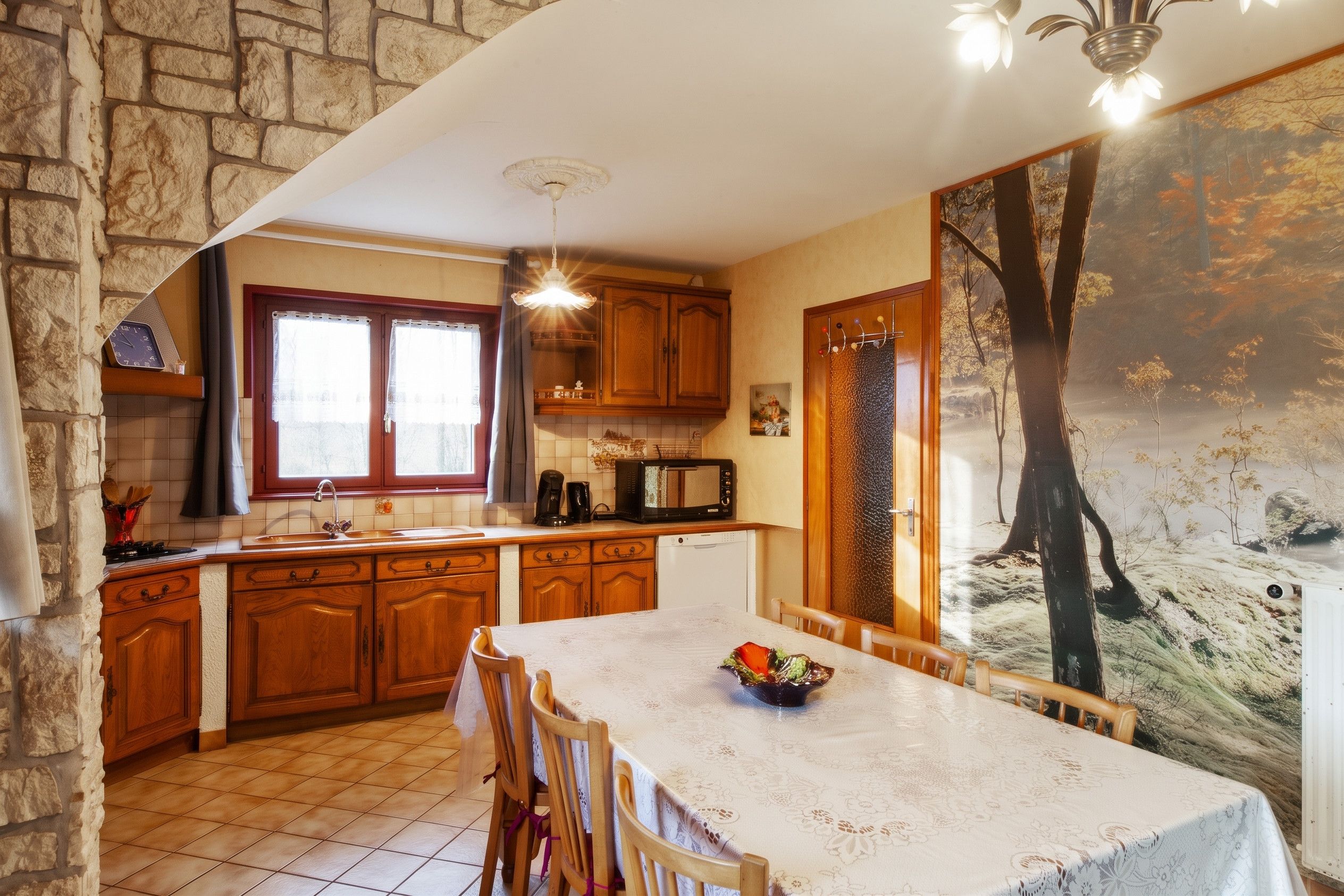 Une cuisine avec des meubles de rangement en bois, une table à manger avec des chaises, un four à micro-ondes et une grande fenêtre.