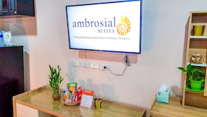A reception area with a sign for Ambrosial Suites, a glass table with snacks, a potted plant, and a small shelf with decorative items. - Walking Street Residence - Boutique Suites (Pattaya)