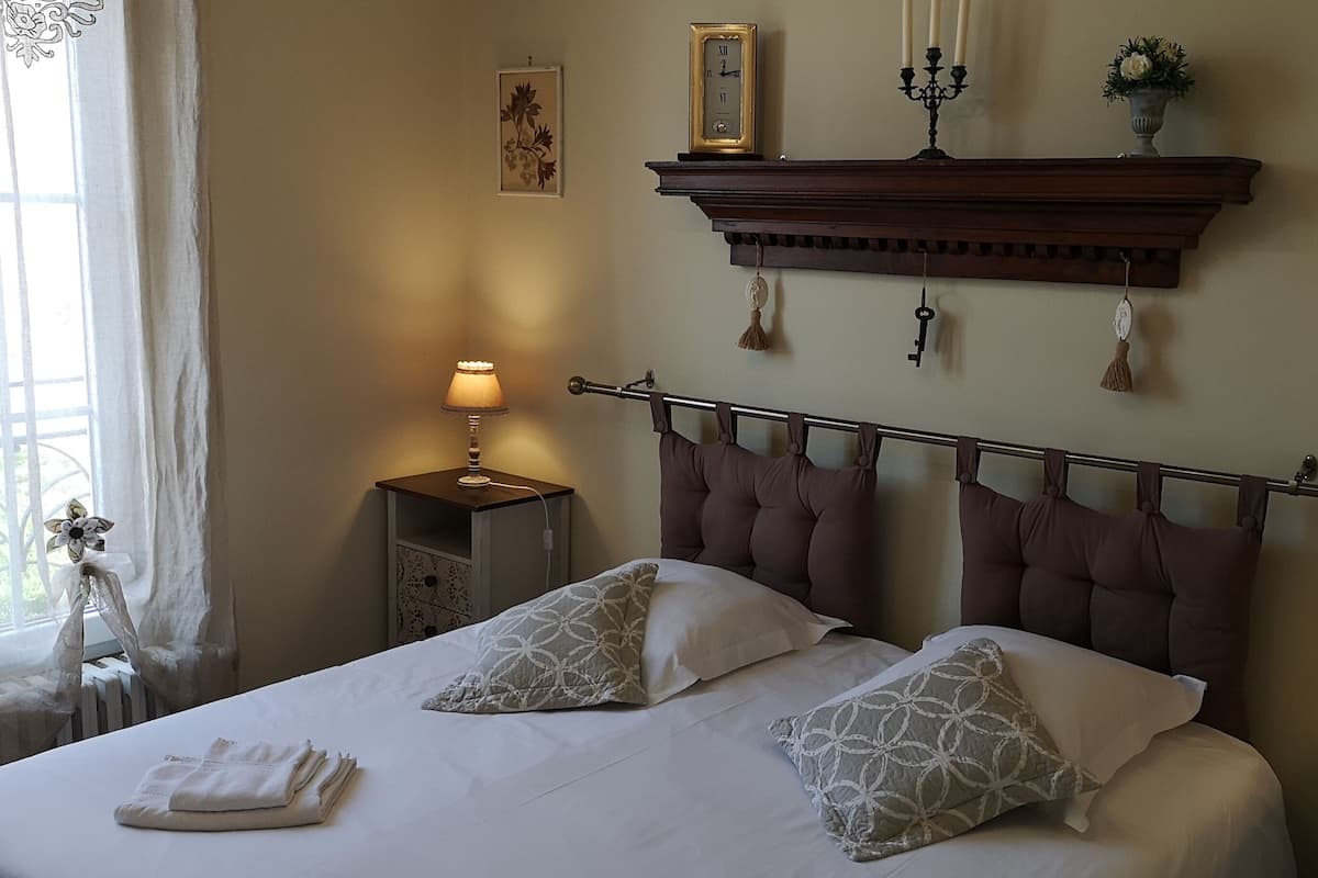 A neatly made bed with two pillows, a bedside table with a lamp, a clock on the wall, and a radiator.