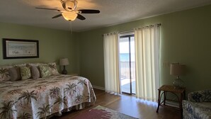 A bedroom with a bed, a ceiling fan, a nightstand, a lamp, a chair, a window with curtains, and a view of the ocean. - 213 Seashack- Your Dream Beach Retreat, Steps from the Shore (St Joe Beach)