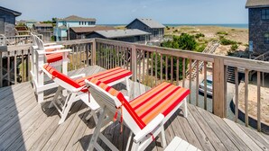 Een houten terras met witte stoelen en rode kussens, met uitzicht op een strand en huizen.