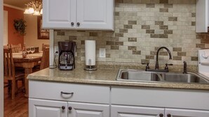 Una cocina con armarios blancos, salpicadero de azulejos, un lavamanos doble y una cafetera sobre la encimera.