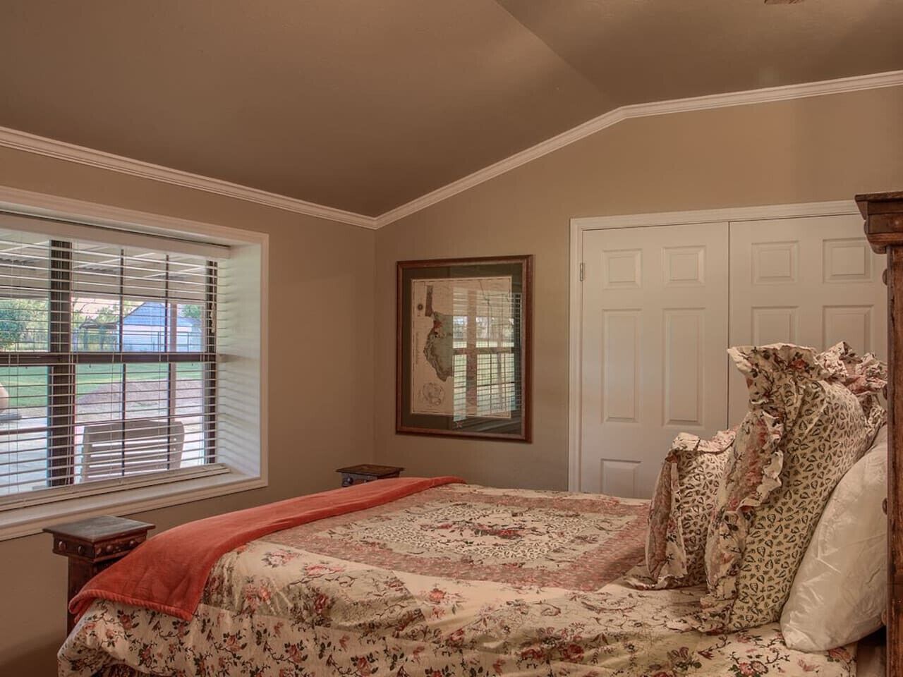 A bedroom with a bed, a window with blinds, a framed picture on the wall, and a door.