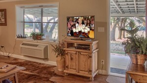 Una sala de estar con televisor sobre un mueble de madera, una ventana con persianas y una puerta que da al exterior.