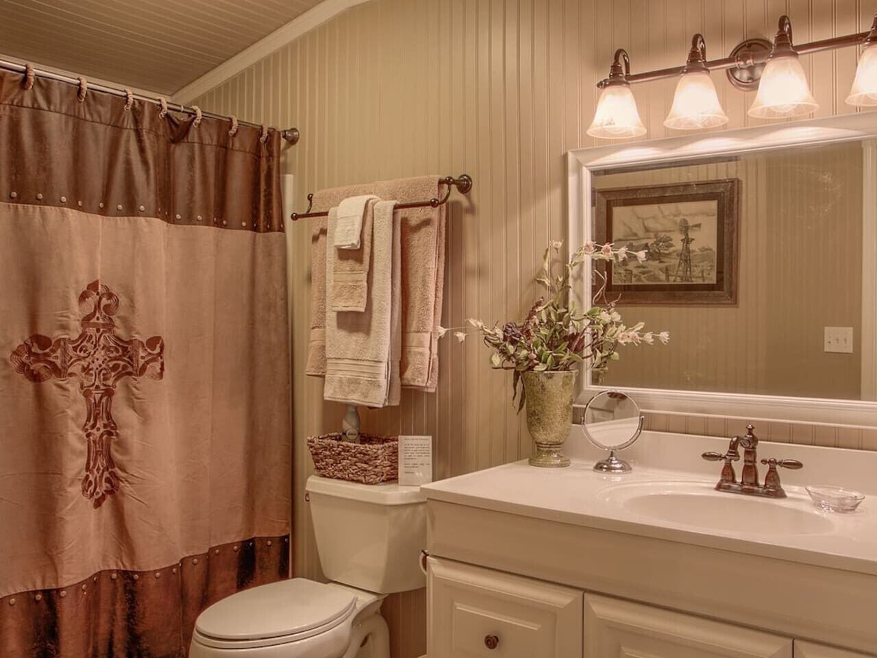 Un baño con cortina de ducha con diseño de cruz, un inodoro, un lavabo con un florero y un espejo con un cuadro enmarcado.