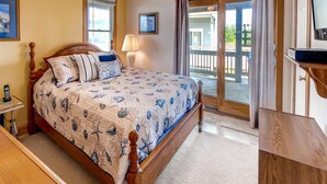 A bedroom with a bed, a bedside table, a lamp, and a view of the outside balcony. - Oceanview Home w/ Private Pool, Hot Tub, and Game Room. It's Simply Irresistible (Waves)