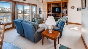 A living room with a sofa, armchairs, a coffee table, and a TV. There is a bookshelf with various items and a window with a view of houses. - Oceanview Home w/ Private Pool, Hot Tub, and Game Room. It's Simply Irresistible (Waves)
