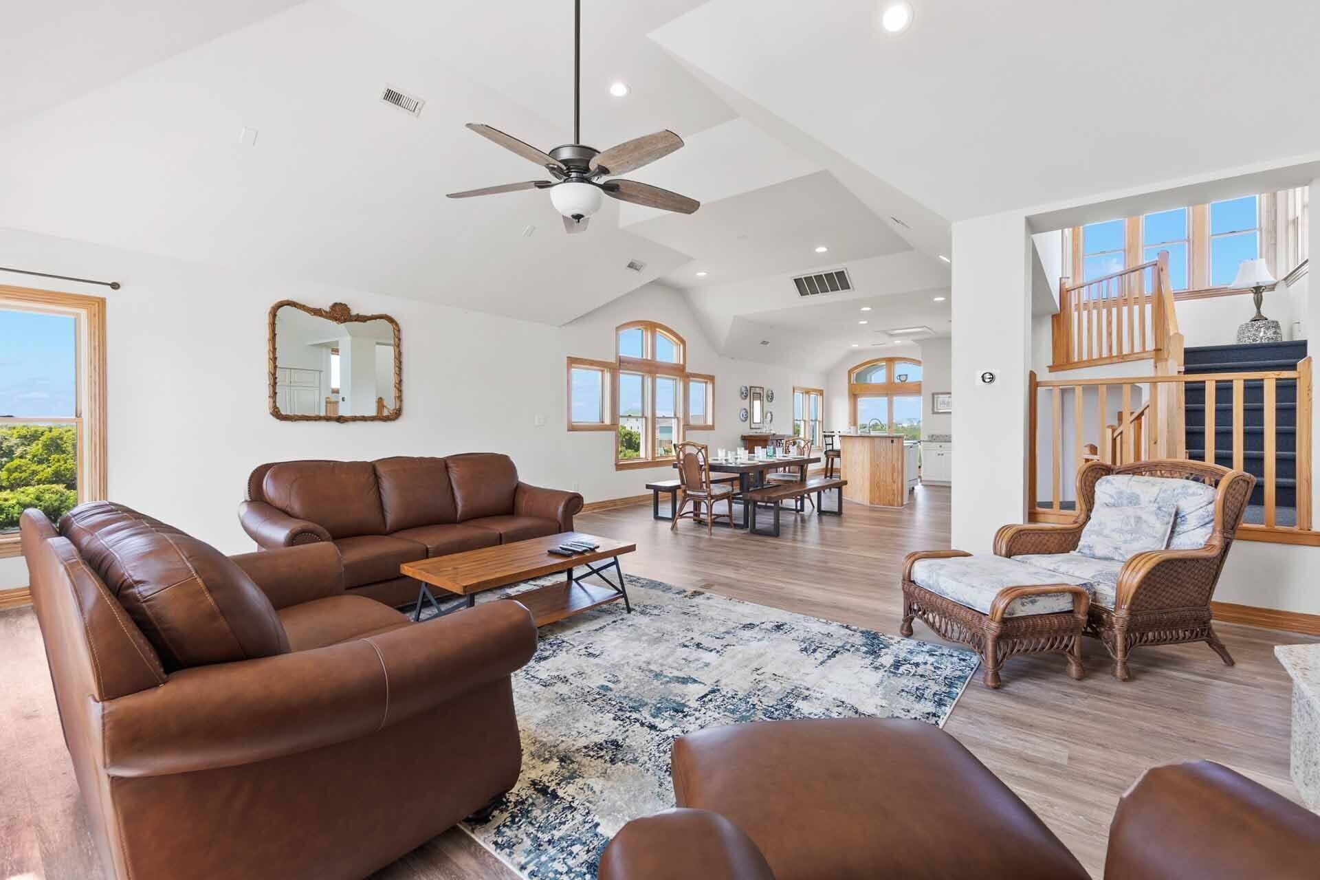 A spacious living room with a brown leather sofa, a dining area with wooden furniture, and a view of the outdoors through large windows.