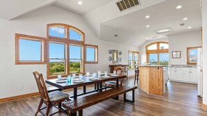 Un comedor amplio con una mesa y bancos de madera, ventanas grandes con vistas, y una cocina con armarios blancos.