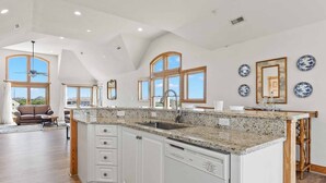 Una cocina amplia con armarios blancos, encimeras de granito y una isla grande. Hay varias ventanas que ofrecen luz natural y vistas al exterior.