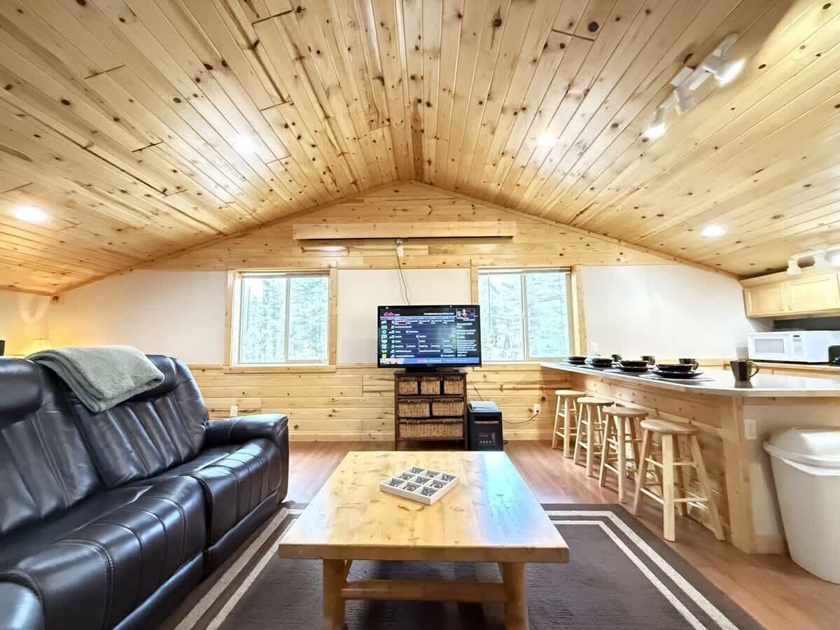 Ein gemütliches Blockhaus-Interieur mit Holzbalkendecke, einer schwarzen Ledersofa, einem Holztisch, einem Fernseher und einer Küchenzeile mit Barhocker.