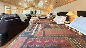 A room with a wooden ceiling, a large bed with patterned bedding, a black leather sofa, and a nightstand with a lamp.