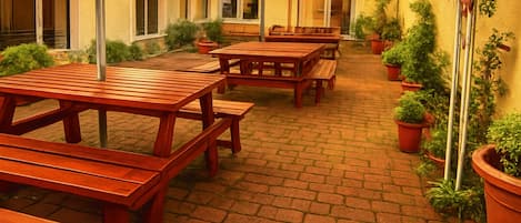 An outdoor patio area with wooden tables and benches, potted plants, and a blue umbrella.