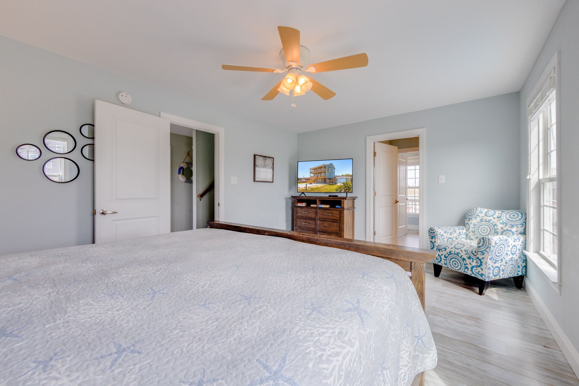 Une chambre avec un grand lit, un ventilateur de plafond, une télévision, une commode, une chaise et une vue sur une maison par la fenêtre.