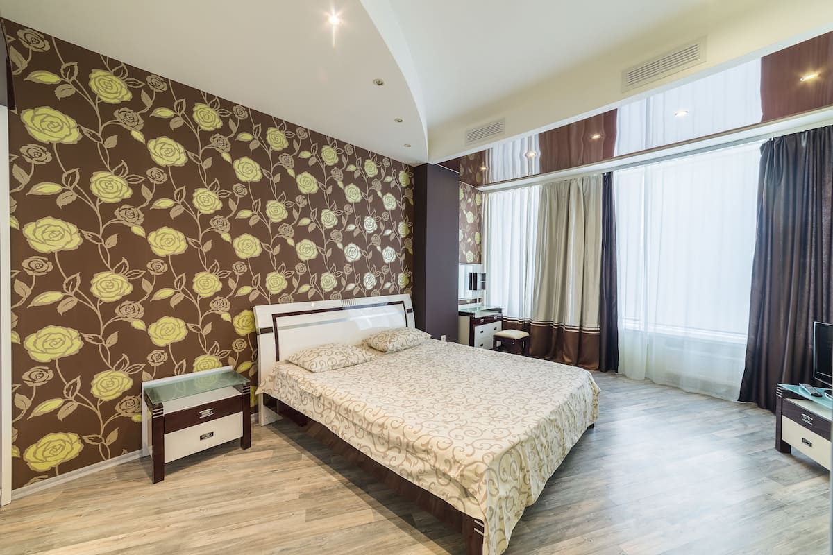A bedroom with a large bed, a bedside table, a desk, and a chair. There is a window with curtains and a view of the outside.