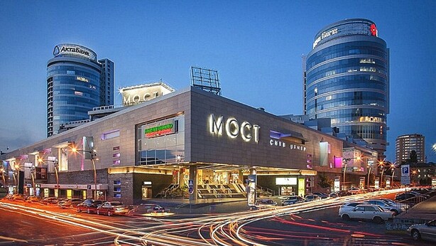 A modern building with the sign "MOCT" prominently displayed, surrounded by other commercial buildings and a busy street with light trails from moving vehicles. - Most City Apartments (Dnipro)