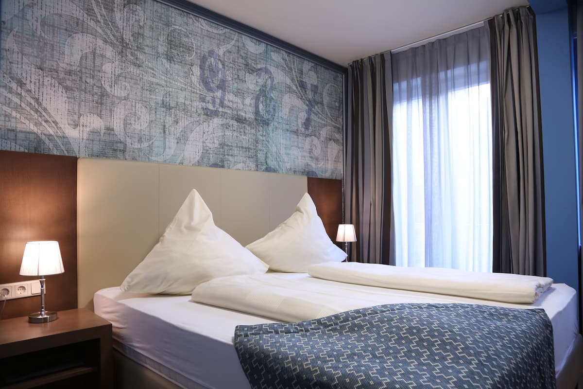 A hotel room with a large bed, two bedside lamps, a headboard with a decorative panel, and a window with curtains.