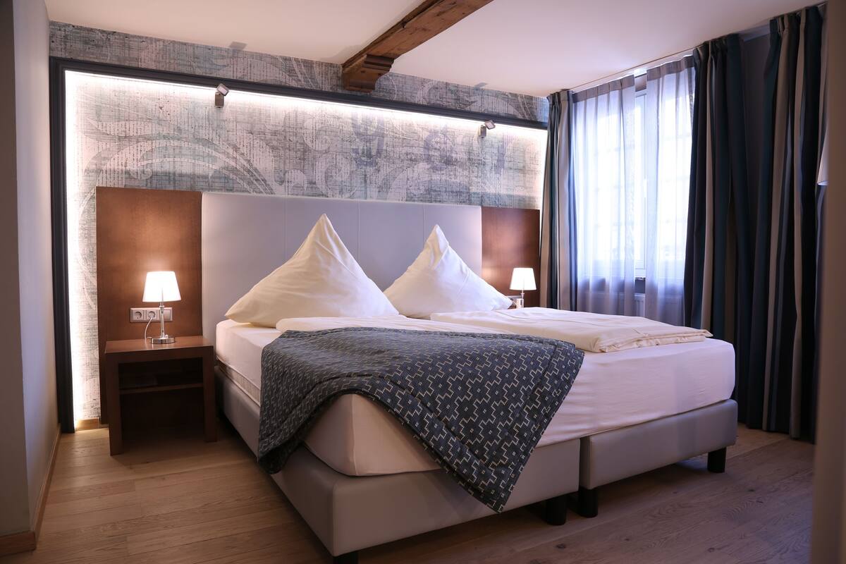 A modern hotel room with a large bed, two bedside tables with lamps, a headboard with a light panel, and a view of the outdoors through sheer curtains.