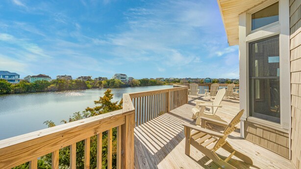 A wooden deck with chairs and a table overlooking a calm body of water, with houses in the background. - Oceanview Dog Friendly Home w/ Private Pool, Hot Tub, Game Room, Community Perks (Waves)
