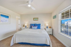 Une chambre à coucher comprenant un lit, deux tables de chevet, un ventilateur de plafond, une fenêtre avec vue et un tableau encadré accroché au mur.