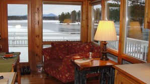 A cozy cabin interior with a wooden floor, a floral sofa, a table with a lamp, and large windows offering a view of a snowy landscape. - AMAZING WATERFRONT SUNSETS ! (Ossipee)