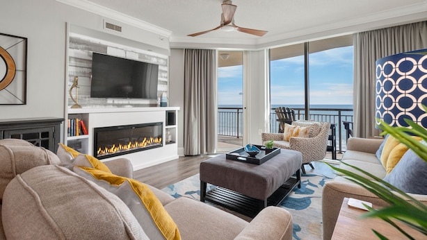 A modern living room with a fireplace, comfortable seating, a television, and a balcony with a view of the ocean.