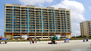 A large hotel with multiple balconies, a sandy beach in front, and umbrellas.