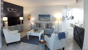 Una sala de estar con un sofá, sillones, una mesa de centro y un televisor. Hay un cuadro en la pared y una lámpara sobre una mesita de noche.