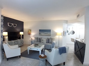 A living room with a sofa, armchairs, a coffee table, and a TV. There is a painting on the wall and a lamp on a side table.