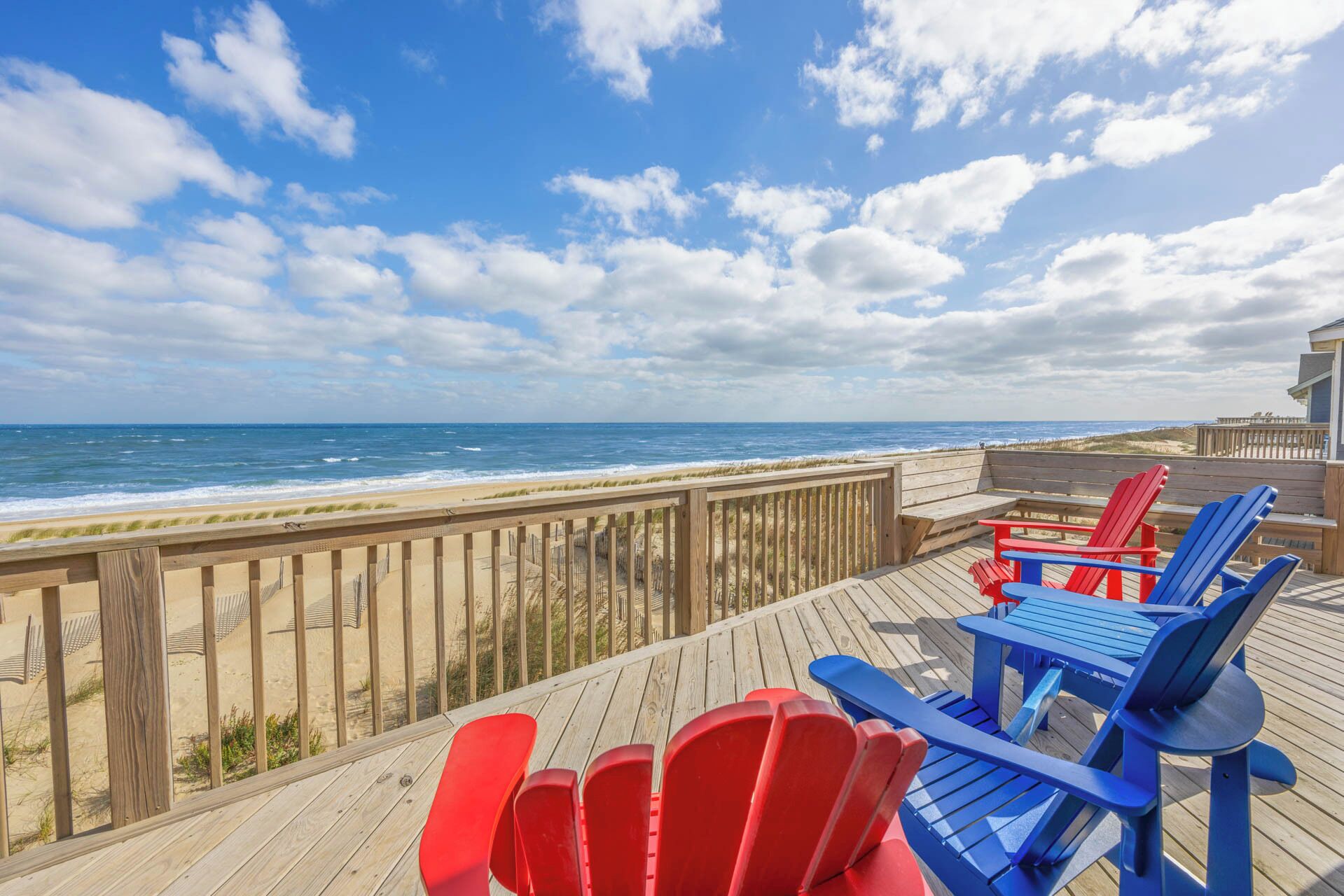A wooden deck with red and blue Adirondack chairs overlooking a beach with a wooden railing.