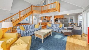 A spacious living area with a high ceiling, a wooden staircase, and a kitchen in the background. - Oceanfront Dog Friendly Home w/ Hot Tub, Foosball, and Direct Beach Access (Avon)