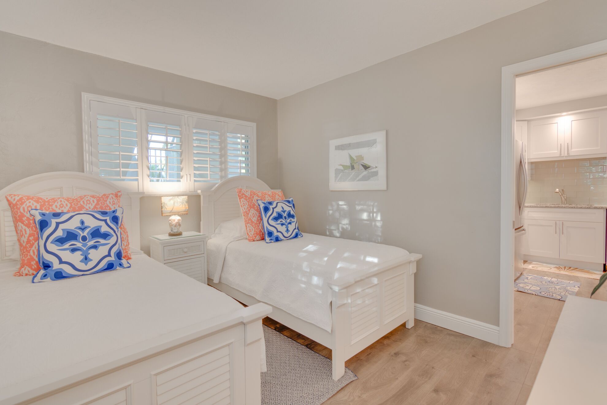 A bedroom with two beds, a window with shutters, a nightstand with a lamp, and a kitchen visible through an open door.