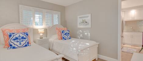 A bedroom with two beds, a window with shutters, a nightstand with a lamp, and a kitchen visible through an open door.