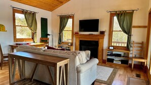 Una sala de estar acogedora con un sofá, una mesa de centro de madera, chimenea y ventanas amplias con cortinas.